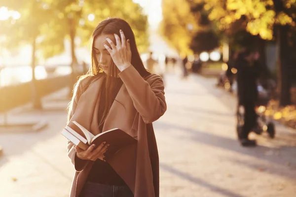 一个年轻漂亮的黑发姑娘正在看一本棕色的书 她用左手整理着头发 站在秋天的公园里 身穿一件休闲装 背景模糊 复制着一片空白 — 图库照片