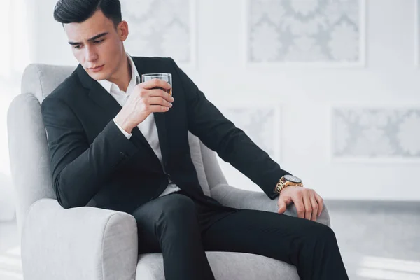 Young elegant guy in black suit sitting on white chair and holds glass with alcohol.