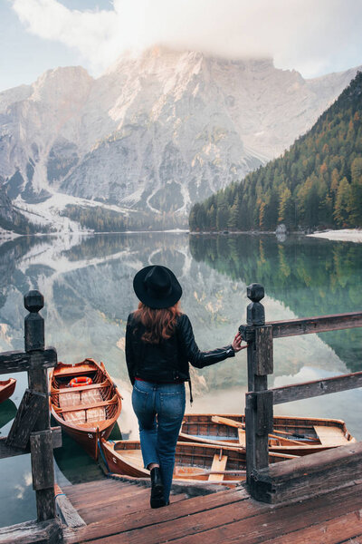 Holds on a wooden pier. Woman in black hat enjoying majestic mountain landscape near the lake with boats.