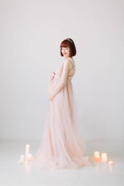 Katta, tam uzunlukta dekoratif yanan mumlar ile beyaz arka plan üzerinde ayakta uzun pembe elbiseli güzel hamile kadın