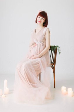 beyaz zemin üzerinde mumlar yanan ile dekore edilmiş sandalye üzerinde oturan pembe içinde oldukça hamile kadın tam uzunlukta