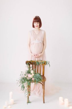Çiçekler beyaz zemin üzerinde mumlar yanan ile dekore edilmiş sandalye yanında ayakta tutan pembe içinde oldukça hamile kadın tam uzunlukta