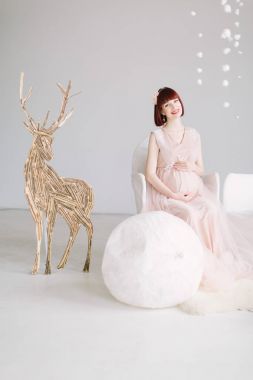 Ahşap geyik figürü ile kış arka plan üzerinde beyaz sandalye üzerinde oturan genç hamile kadın