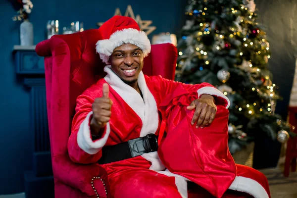 Navidad y Feliz Año Nuevo concepto. Santa Claus africano sonriente sosteniendo un saco rojo con regalos de Navidad mientras está sentado en una silla roja cerca del árbol de Navidad — Foto de Stock