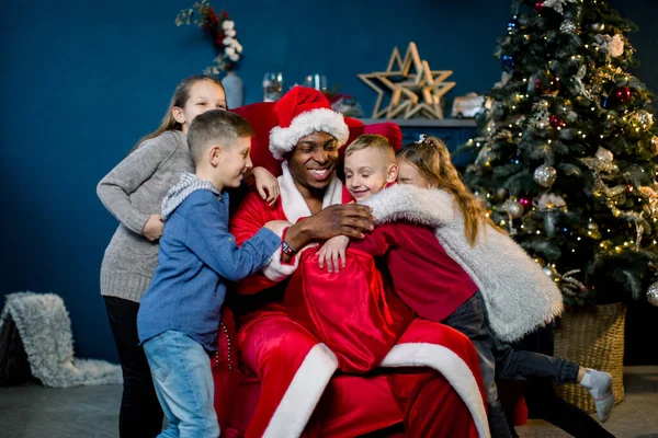 Gelukkig lachen kinderen knuffelen Afrikaanse kerstman zitten op een rode stoel op de achtergrond van een kerstboom en decoraties. — Stockfoto