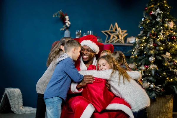 Gelukkig lachen kinderen knuffelen Afrikaanse kerstman zitten op een rode stoel op de achtergrond van een kerstboom en decoraties. — Stockfoto
