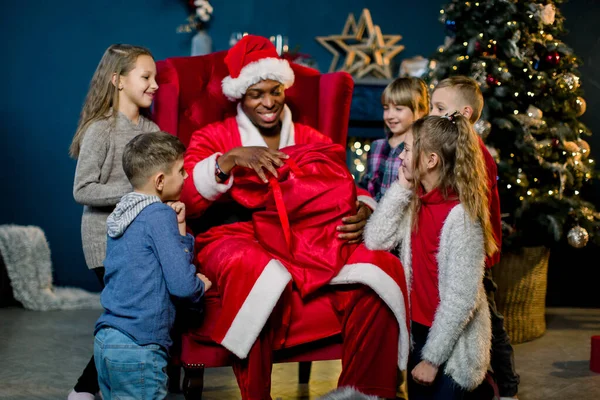 Afrikaanse Kerstman zit in een fauteuil en geeft de cadeautjes aan mooie kleine kinderen in een kerstversierde kamer. Nieuwjaar, winterholidys, Kerstconcept — Stockfoto