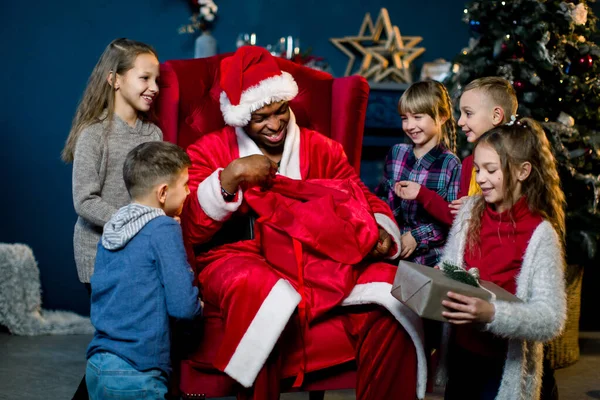 Babbo Natale africano seduto in poltrona e dare i regali ai bellissimi bambini piccoli in una stanza decorata di Natale. Capodanno, vacanze invernali, concetto di Natale — Foto Stock