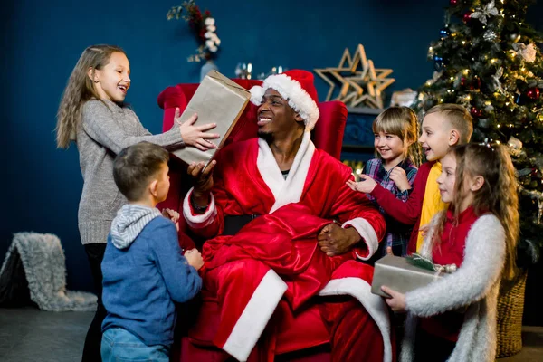 Afrikaanse Kerstman zit in een fauteuil en geeft de cadeautjes aan mooie kleine kinderen in een kerstversierde kamer. Nieuwjaar, winterholidys, Kerstconcept — Stockfoto