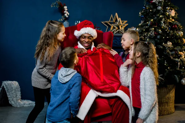 Afrikaanse Kerstman zit en geeft cadeautjes aan mooie kleine kinderen in een kerstversierde kamer. Afrikaanse kerstman en gelukkige kleine kinderen op de achtergrond van de kerstboom — Stockfoto