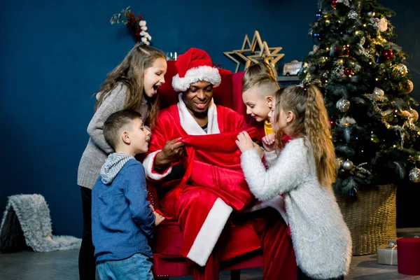 Vijf kinderen kijken in de zak van de Afrikaanse Kerstman en lachen, Afrikaanse Kerstman zit en geeft cadeautjes aan mooie kleine kinderen in een kerstversierde kamer. — Stockfoto