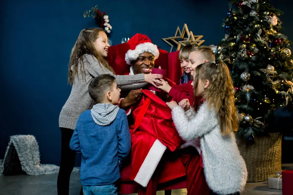 Vijf kinderen kijken in de zak van de Afrikaanse Kerstman en lachen, Afrikaanse Kerstman zit en geeft cadeautjes aan mooie kleine kinderen in een kerstversierde kamer. — Stockfoto