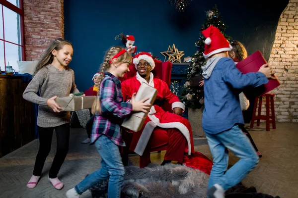 De Afrikaanse Kerstman zit in een rode fauteuil. Gelukkige kinderen rennen met de kerstman geschenken in de kerstkamer — Stockfoto