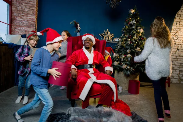 De Afrikaanse Kerstman zit in een rode fauteuil. Gelukkige kinderen rennen met de kerstman geschenken in de kerstkamer — Stockfoto