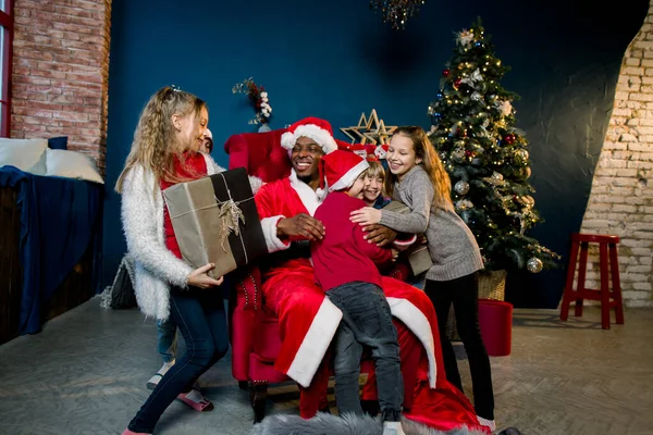 Babbo Natale africano si siede su uno sfondo di un albero di Natale e presenta regali ai bambini — Foto Stock
