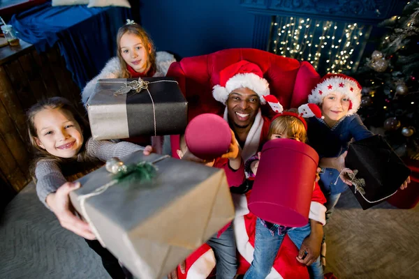 Beaux enfants avec des cadeaux dans leurs mains autour du Père Noël africain assis dans un fauteuil sur le fond d'un arbre de Noël . — Photo