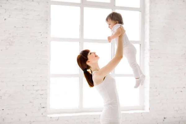 Felice affascinante madre sportiva in bianco indossare tenendo carina bambina in alto sopra la testa, mentre in piedi contro la grande finestra in palestra sala luce — Foto Stock