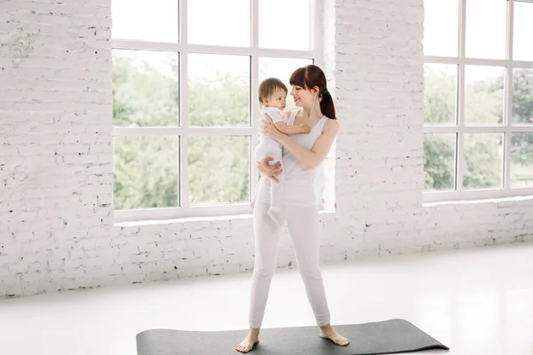 Portrait of beautiful young mom in sports wear holding her charming little baby, standing on black yoga mat, looking at baby and smiling while standing against window