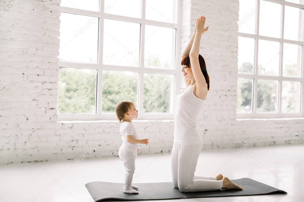 sports young mother in white wear is engaged in fitness and yoga with a baby girl at light gym with big windows