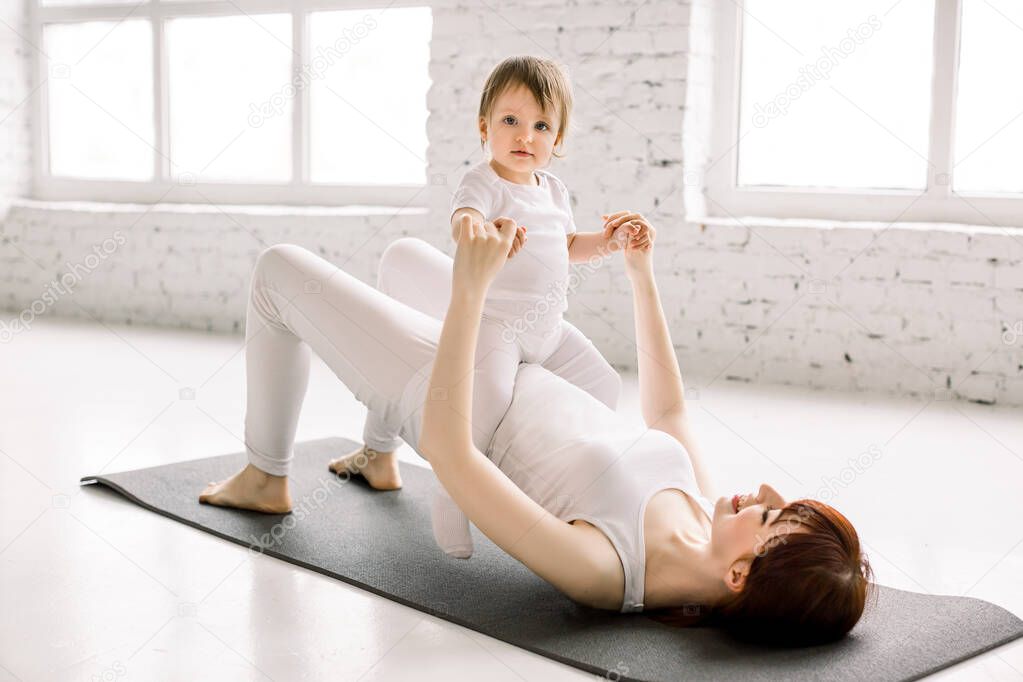Young smiling mother doing yoga exercise at gym, wearing white sportswear, together with funny little daughter, enjoying activities with baby, fun and sport practice. Healthy lifestyle concept photo