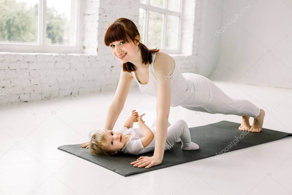 Portrait of beautiful young mother in white sports wear with her charming little baby girl doing exercises on the black mat in training session at gym