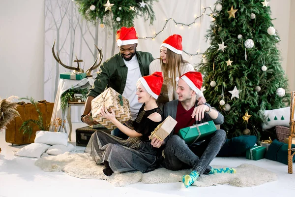 Gruppe junger multiethnischer Freunde in der Nähe des schön geschmückten Weihnachtsbaums, tauschen Weihnachtsgeschenke aus und haben Spaß. — Stockfoto