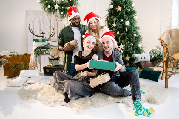 새해, 크리스마스, 겨울 휴일 컨셉트. 산타 모자를 쓴 젊은이들 이 바닥에 앉아 크리스마스를 축하하며 현재의 상자를 들고 즐거운 시간을 보내고 있습니다.. — 스톡 사진