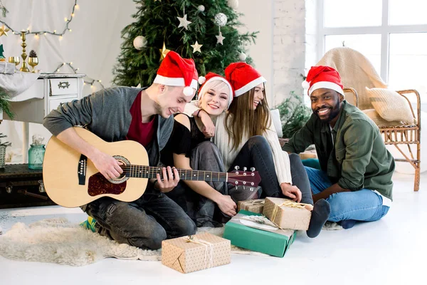 Fröhliche junge vier internationale Menschen feiern gemeinsam Weihnachten und Neujahr in einem gemütlich dekorierten Studio. weißer Mann spielt Gitarre. Winterferienkonzept. — Stockfoto