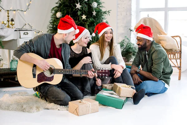 Fröhliche junge vier internationale Menschen feiern gemeinsam Weihnachten und Neujahr in einem gemütlich dekorierten Studio. weißer Mann spielt Gitarre. Winterferienkonzept. — Stockfoto