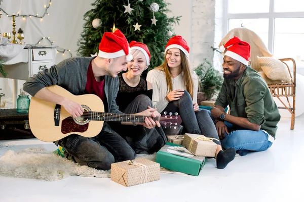 Das Bild zeigt eine Gruppe von vier Freunden, die zu Hause Weihnachten feiern. junger kaukasischer Mann spielt Gitarre und die Mädchen und der afrikanische Mann lächeln und singen Weihnachtslieder — Stockfoto