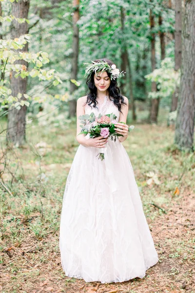 La novia con corona en la cabeza en un elegante vestido de novia en el bosque natural con un ramo rústico. Sesión de fotos en un bosque de pinos al estilo de las bellas artes . —  Fotos de Stock