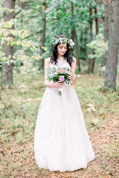 Hermosa novia con corona de eucalipto en la cabeza de pie en el bosque y mirando ramo de boda de diferentes flores. Estilo rústico —  Fotos de Stock