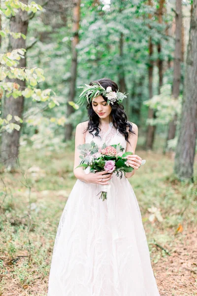 Hermosa novia con corona de eucalipto en la cabeza de pie en el bosque y mirando ramo de boda de diferentes flores. Estilo rústico —  Fotos de Stock