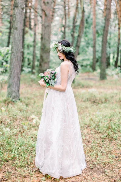 La vue arrière de la charmante mariée brune en couronne de verdure sur la tête tenant le bouquet de mariage pendant la promenade dans la forêt . — Photo