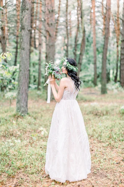 La vue arrière de la charmante mariée brune en couronne de verdure sur la tête tenant le bouquet de mariage pendant la promenade dans la forêt . — Photo