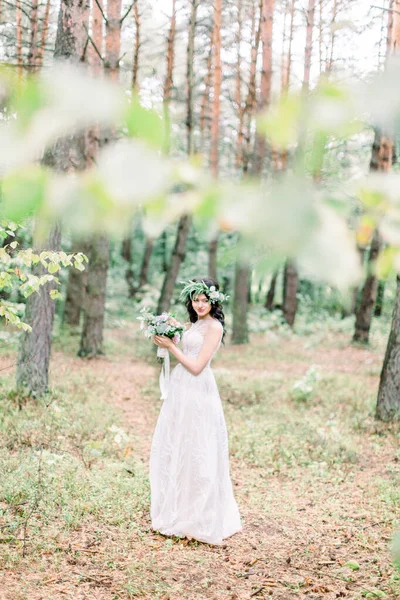 La mariée en robe volante marchant dans la forêt de pins. Mariée en couronne sur la tête tient son bouquet dans les mains et sourit — Photo