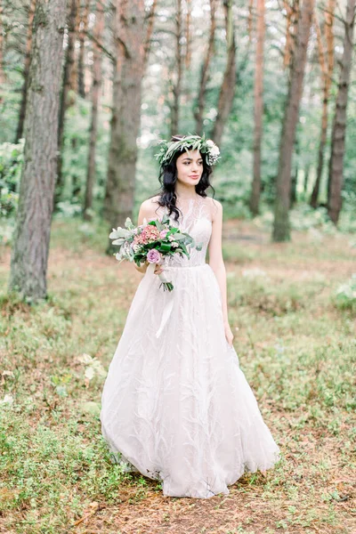 Vue complète de la charmante mariée en couronne de verdure avec le bouquet de mariage regardant de côté dans la forêt de printemps . — Photo