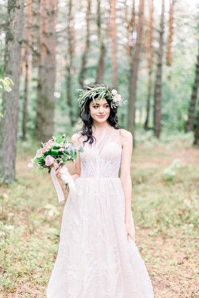 Hermosa novia caminando en un bosque de coníferas en una corona en su cabeza y un vestido de novia de lujo, la celebración de rústico ramo de bodas —  Fotos de Stock