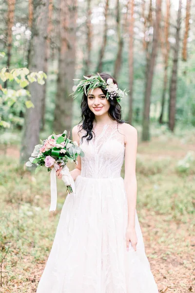 Hermosa novia caminando en un bosque de coníferas en una corona en su cabeza y un vestido de novia de lujo, la celebración de rústico ramo de bodas —  Fotos de Stock