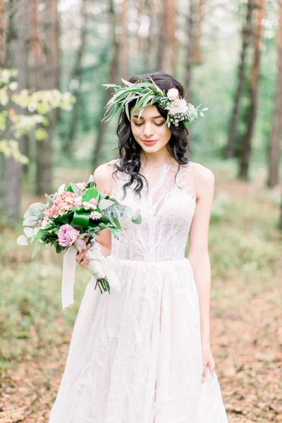 Wedding ceremony in rustic style. Beautiful bride with green wreath on the head, with bouquet in her hand, posing in pine forest