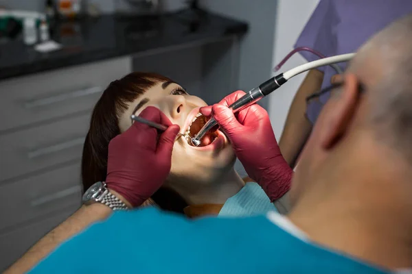 Medical dentist team in modern dental office doing treatment of tooth for female young patient. Senior male dentist and his assistant on the background