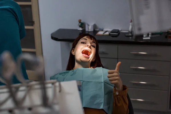 Улыбающаяся молодая женщина, показывающая большой палец, готова к стоматологическому лечению, сидя в стоматологическом кабинете. Улыбающаяся и счастливая молодая женщина после успешного лечения без боли — стоковое фото