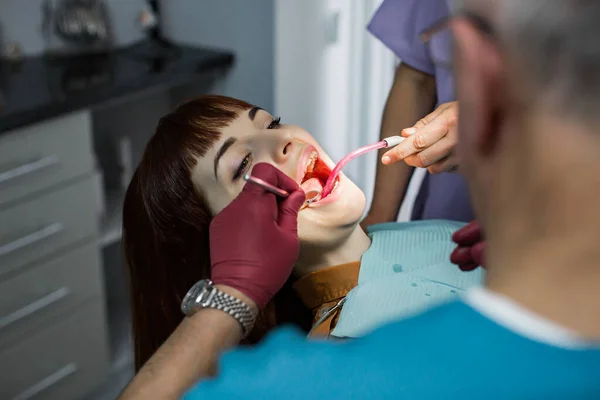 Jonge vrolijke blanke vrouw tijdens het onderzoek in de tandheelkundige kliniek. Senior mannelijke arts tandarts doet tandheelkundige orale check in moderne kliniek — Stockfoto