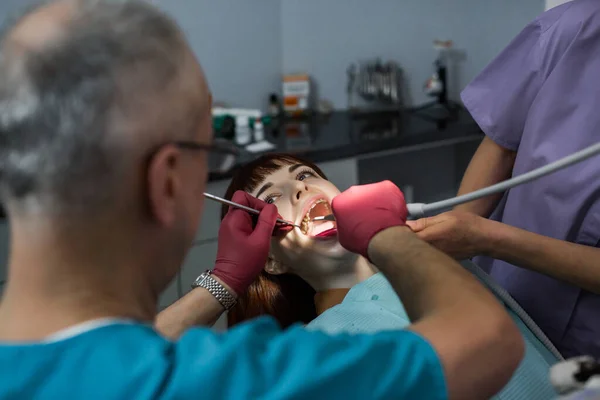 Equipe de dentista médica em consultório odontológico moderno fazendo tratamento de dente para paciente jovem do sexo feminino. Médico dentista sénior e seu assistente em segundo plano — Fotografia de Stock