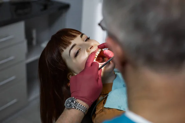 Jonge vrolijke blanke vrouw tijdens het onderzoek in de tandheelkundige kliniek. Senior mannelijke arts tandarts doet tandheelkundige orale check in moderne kliniek — Stockfoto