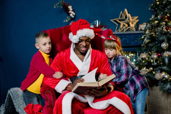 小さな美しい子供たち、女の子と男の子、クリスマスツリーの背景に赤い椅子に座っている間にサンタクロースが読む話を注意深く聞いてください. — ストック写真
