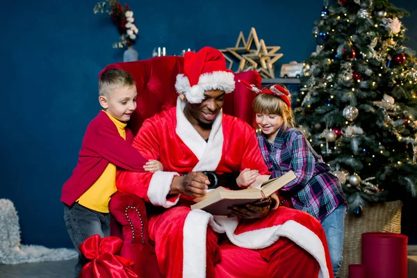 Kleine mooie kinderen, meisje en jongen, luister aandachtig naar het verhaal dat de kerstman leest terwijl hij op een rode stoel zit op de achtergrond van een kerstboom. — Stockfoto