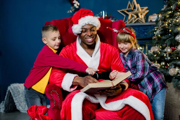 Babbo Natale africano è seduto su una sedia e leggere un libro con storie di Natale per due bambini piccoli — Foto Stock