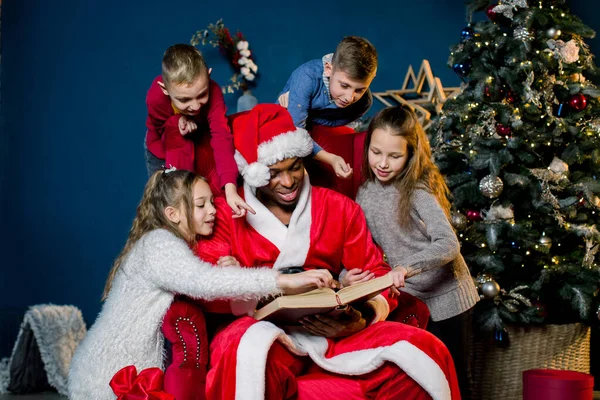La vigilia di Natale, Babbo Natale legge la storia di Natale per i bambini che lo ascoltano da tutte le parti, sullo sfondo di un albero di Natale. . — Foto Stock