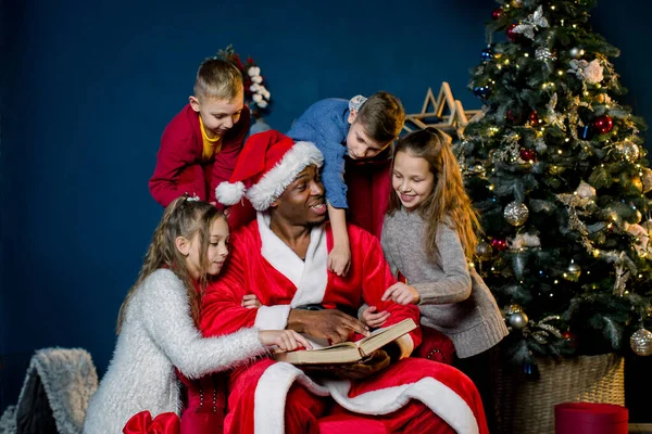 La veille de Noël, le Père Noël lit l'histoire de Noël pour les enfants qui l'écoutent de tous côtés, sur le fond d'un arbre de Noël . — Photo